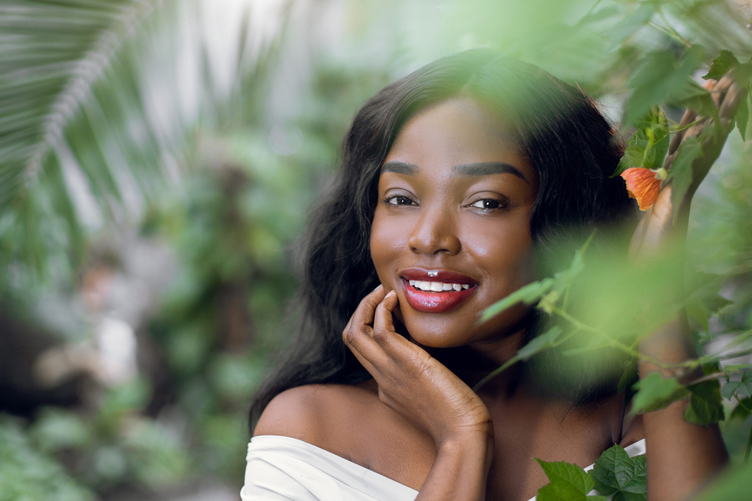 Portrait of exotic charming African dreamy girl, with dark long hair, looking at camera while standing among the leaves of palm and flowering tree. Natural woman beauty. Copy space